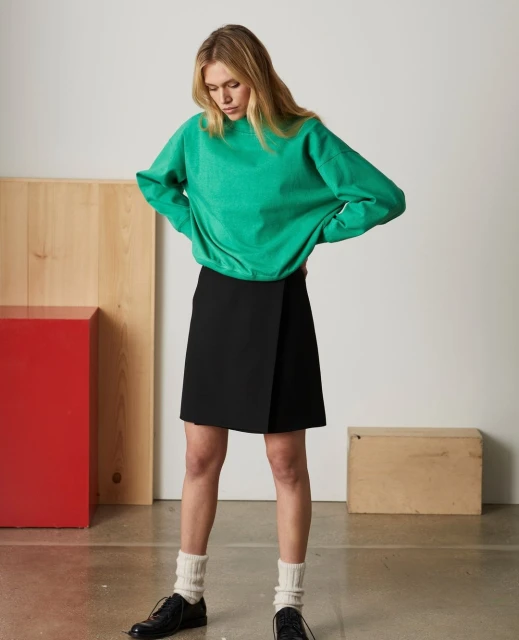 A fair-skinned female fashion model poses in a studio wearing a green sweater, black skirt, white socks, and black dress shoes.