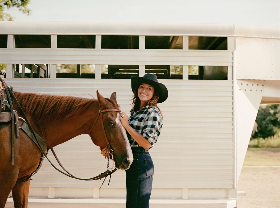 Smiling woman in a cowboy hat stands next to a brown horse with a horse trailer behind them in an outdoor setting on a sunny day.