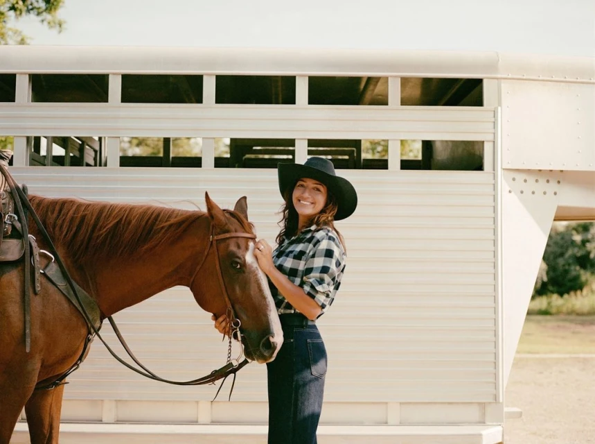 Smiling woman in a cowboy hat stands next to a brown horse with a horse trailer behind them in an outdoor setting on a sunny day.