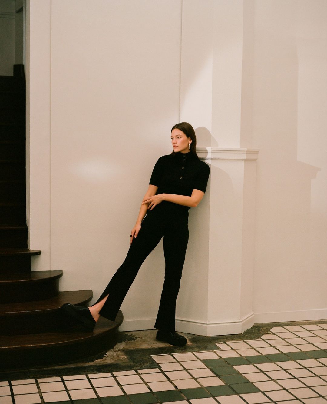 A woman in black leaning against a white wall on a tile floor next to wooden steps, looking away from the camera in an indoor setting.