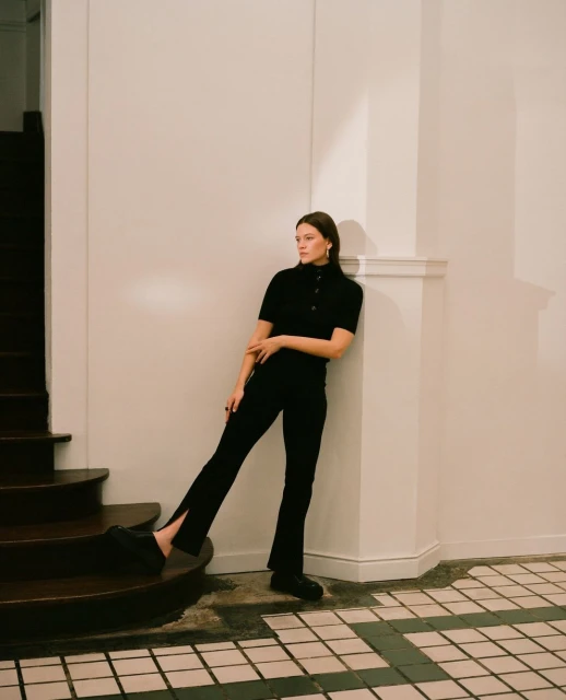 A woman in black leaning against a white wall on a tile floor next to wooden steps, looking away from the camera in an indoor setting.