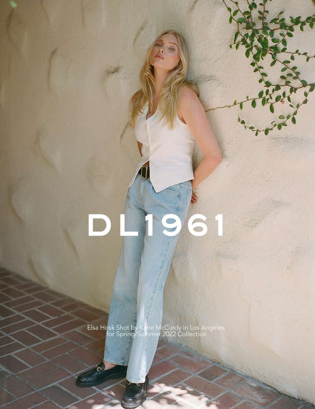 Blonde model Elsa Hosk leans against a textured wall wearing light blue jeans and a white vest top in an outdoor fashion shot.