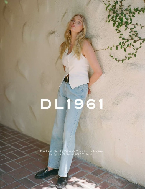 Blonde model Elsa Hosk leans against a textured wall wearing light blue jeans and a white vest top in an outdoor fashion shot.