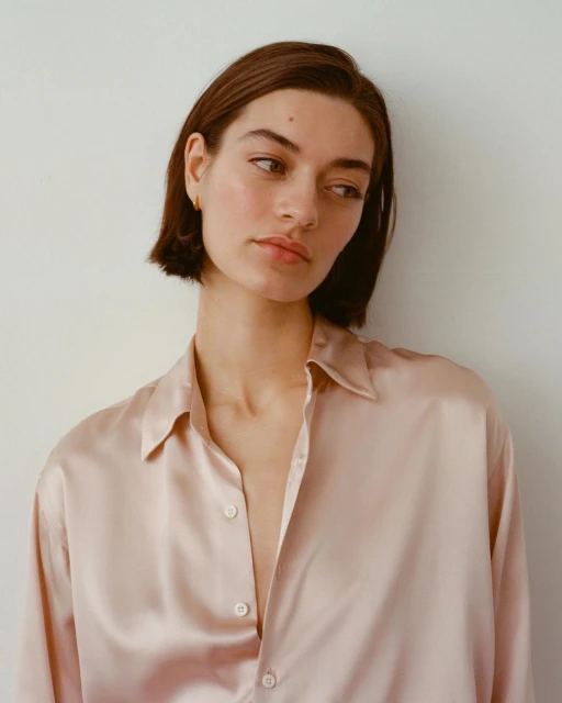 Portrait of a young woman with short brown hair wearing a pink silk shirt against a light background in a natural light setting.