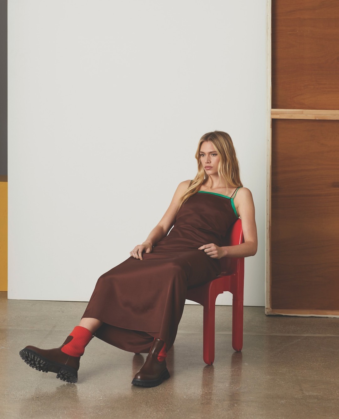 A woman in a slip dress and lug boots sits on a red chair in a photo studio with neutral background panels.