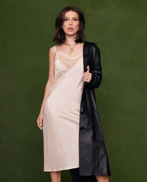 Millie Bobby Brown poses in a studio wearing a cream-colored slip dress with a black coat against a solid green background.