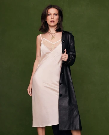 Millie Bobby Brown poses in a studio wearing a cream-colored slip dress with a black coat against a solid green background.