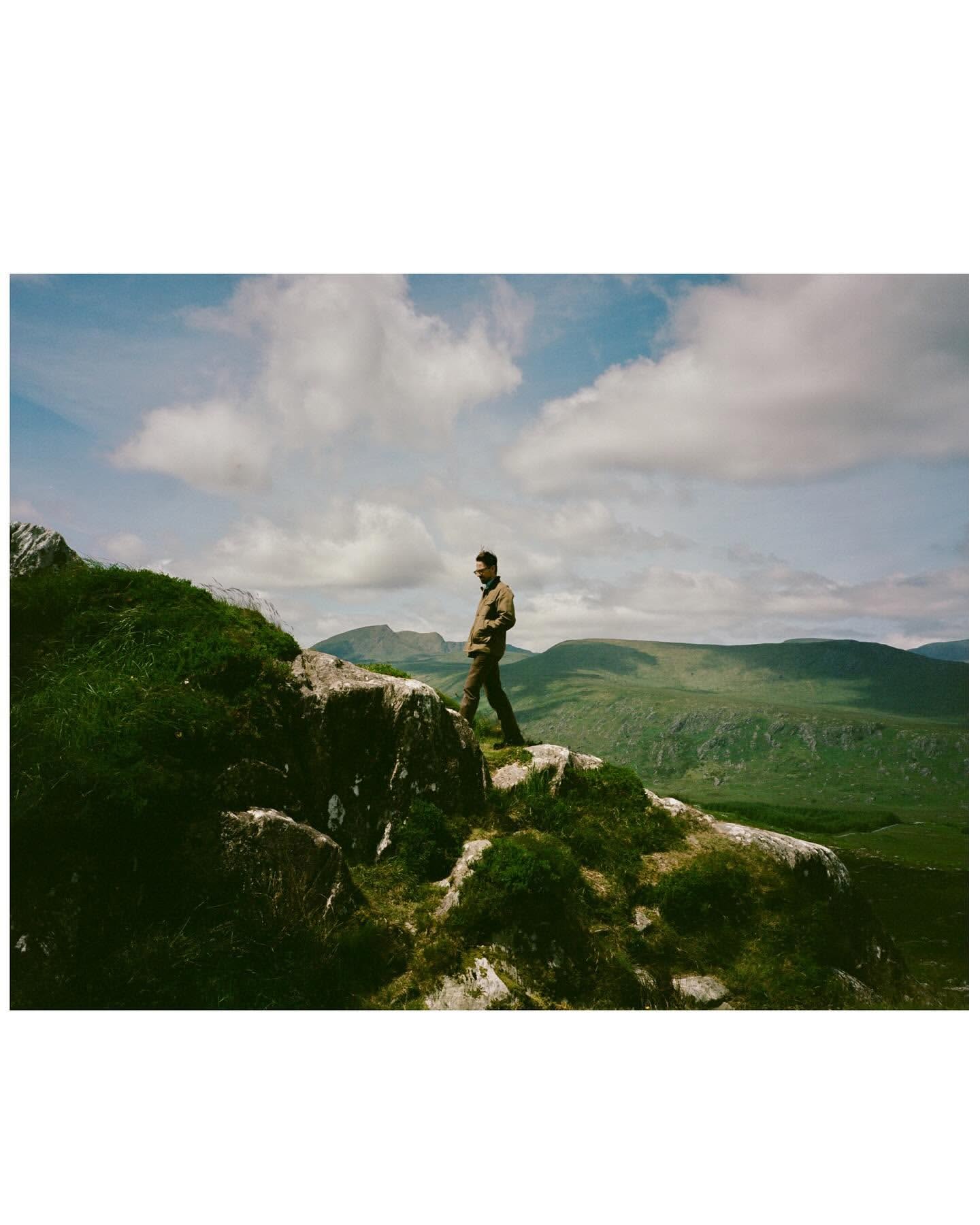 Adult male hiking on a rocky outcrop with green vegetation in a scenic mountain landscape under a blue sky with fluffy white clouds.
