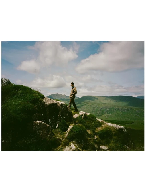 Adult male hiking on a rocky outcrop with green vegetation in a scenic mountain landscape under a blue sky with fluffy white clouds.