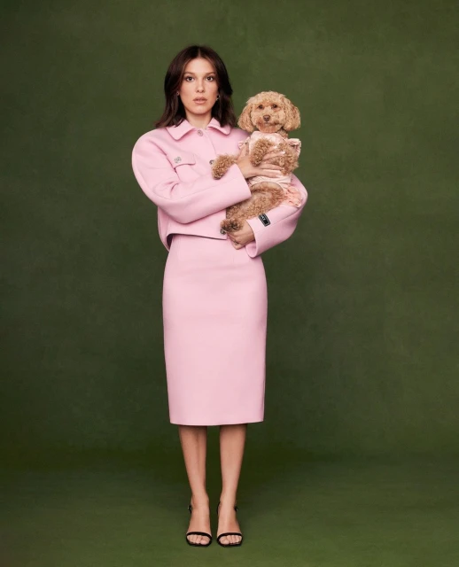 A woman in a pink skirt suit is holding a tan poodle against a green backdrop in a full shot, looking at the camera.