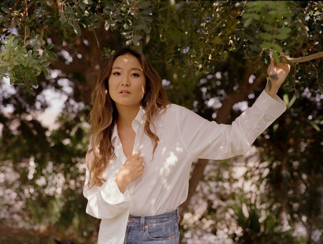 Woman poses outdoors in bright sunlight, wearing a white shirt and jeans, with a backdrop of blurred greenery.