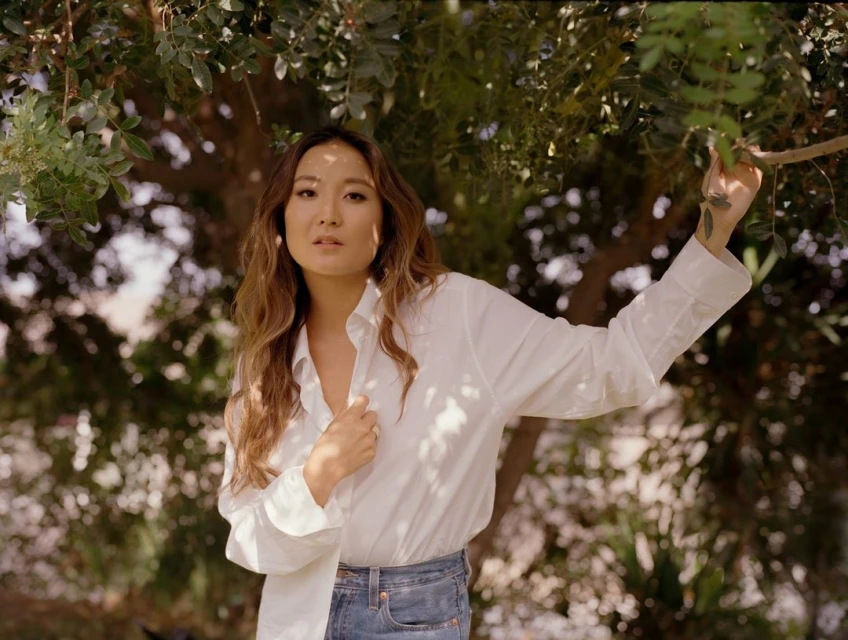 Woman poses outdoors in bright sunlight, wearing a white shirt and jeans, with a backdrop of blurred greenery.