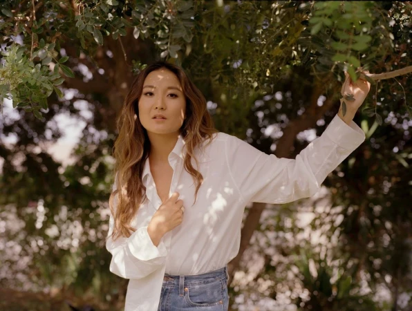Woman poses outdoors in bright sunlight, wearing a white shirt and jeans, with a backdrop of blurred greenery.