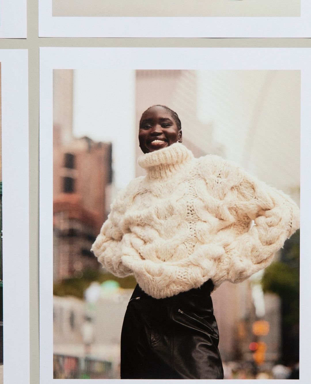 Smiling African woman poses in a knitted sweater and black pants on a city street with blurred buildings in the background in a fashion shot.