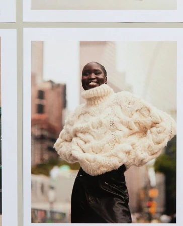 Smiling African woman poses in a knitted sweater and black pants on a city street with blurred buildings in the background in a fashion shot.