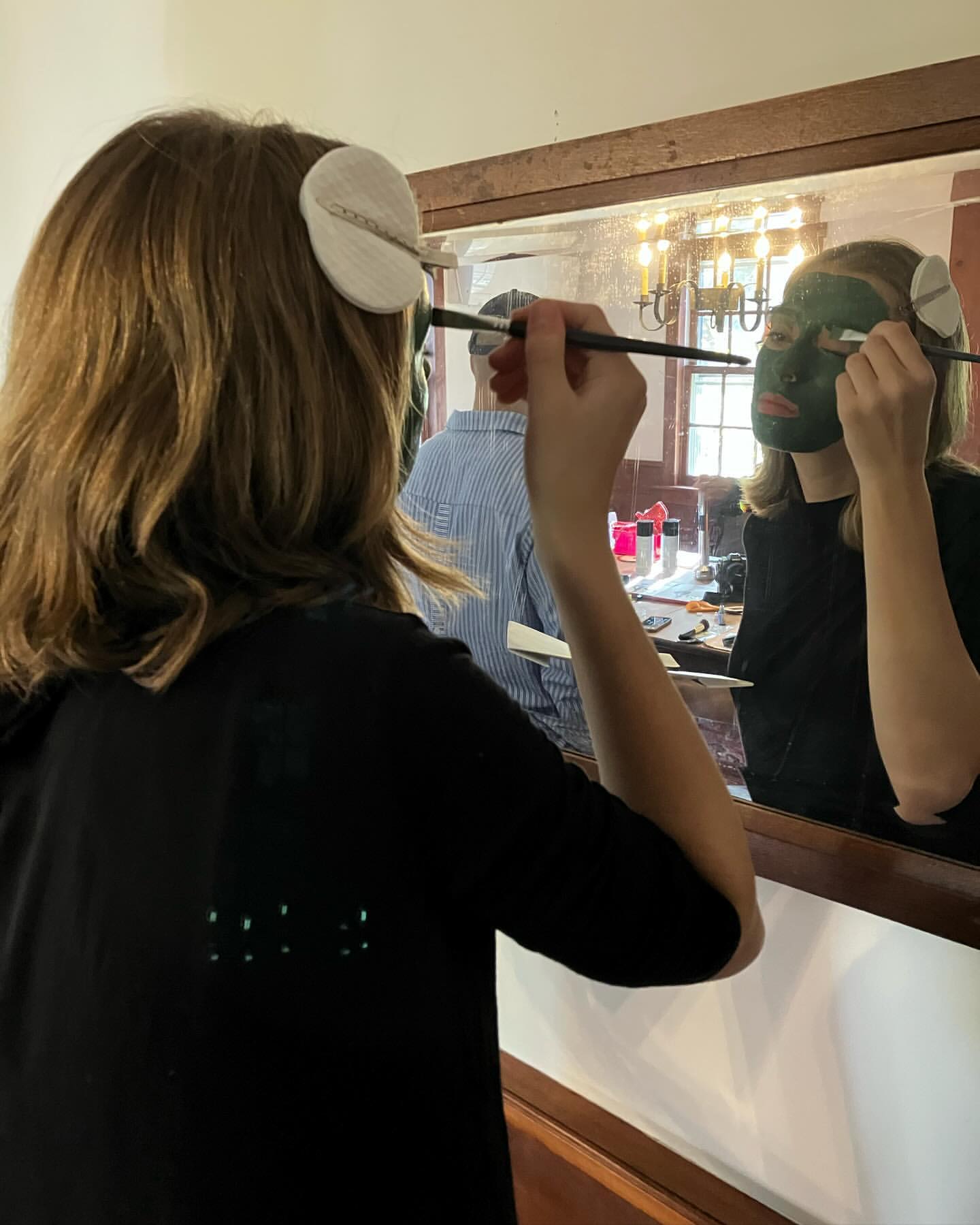 Woman applying a green facial mask in a mirror indoors. Hair is brown and she is wearing a black t-shirt. Vanity is visible.