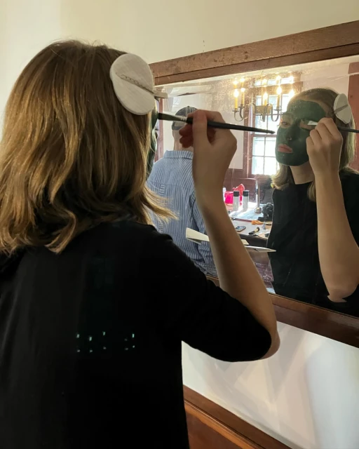Woman applying a green facial mask in a mirror indoors. Hair is brown and she is wearing a black t-shirt. Vanity is visible.