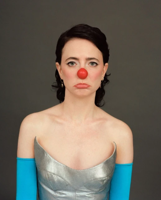 Portrait of a woman with dark hair wearing a silver dress and bright blue sleeves and a red clown nose against a grey background.