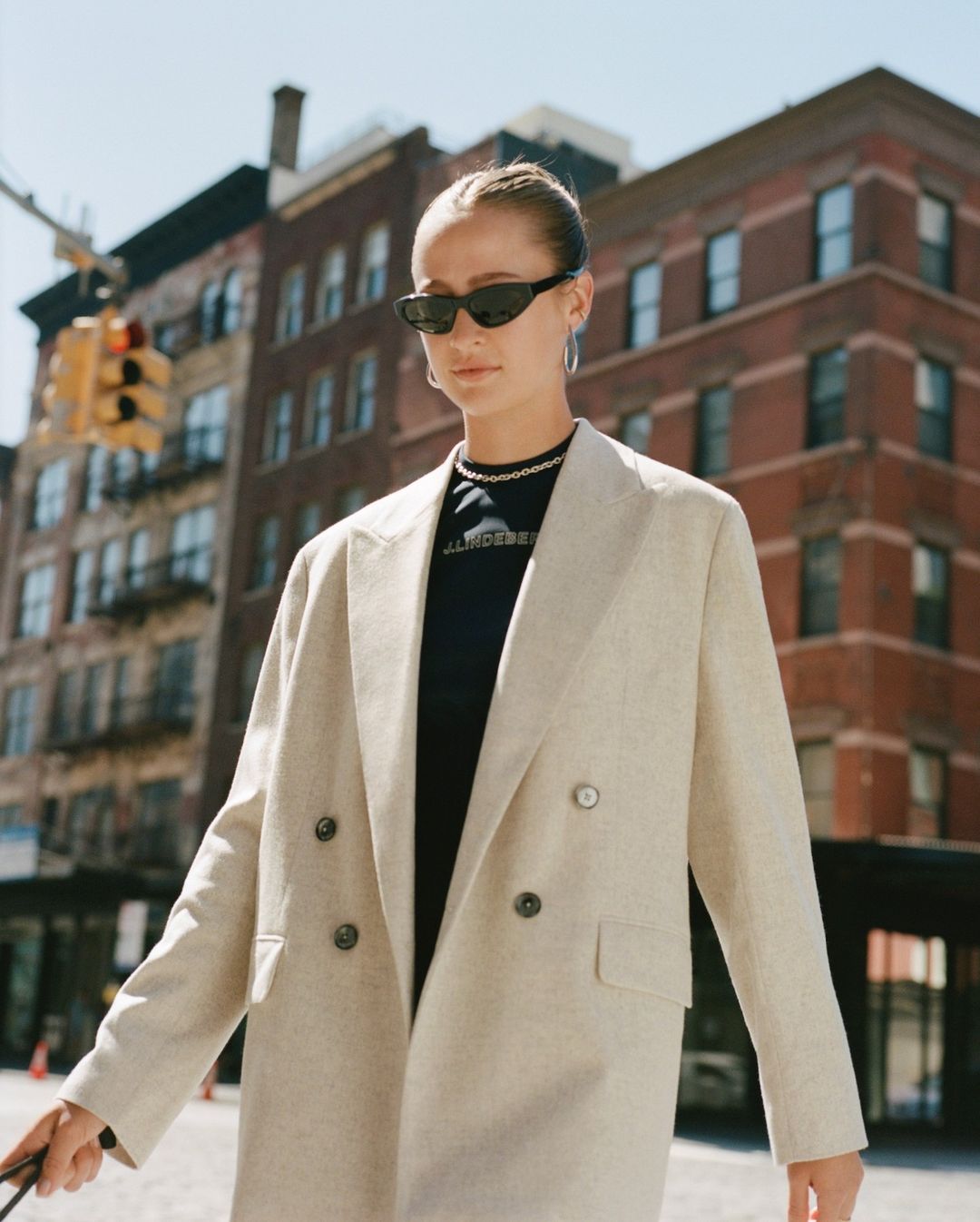 Woman in oversized beige blazer and black sunglasses walking down a city street with red brick buildings in background.