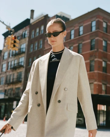 Woman in oversized beige blazer and black sunglasses walking down a city street with red brick buildings in background.