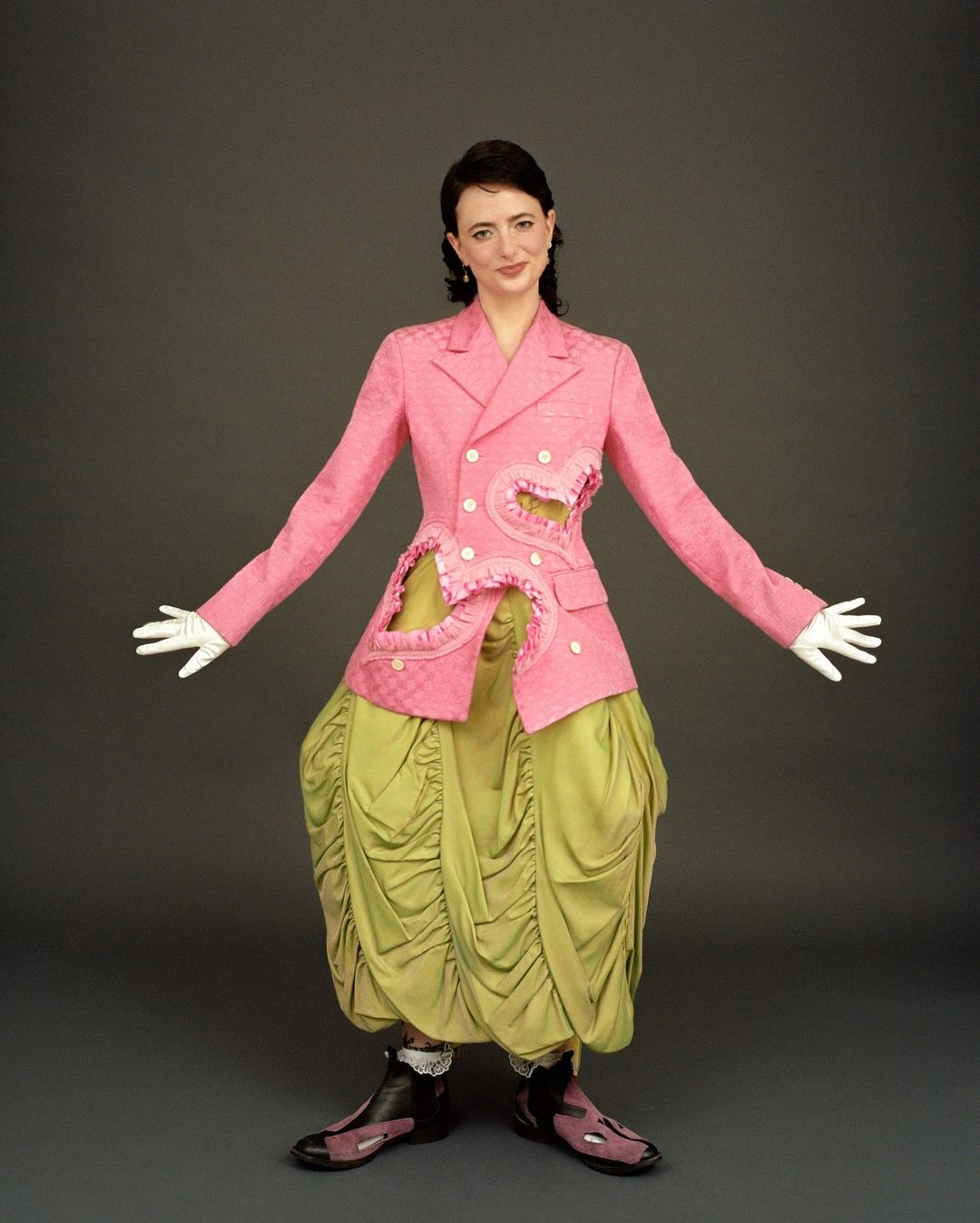 A woman in a pink jacket and green skirt poses in a studio with her arms outstretched against a solid grey background.