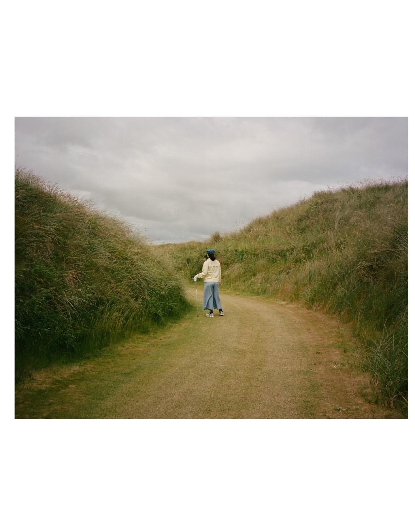A person walks down a grassy path between tall, grassy dunes under a cloudy sky. The person is wearing light-colored clothing.