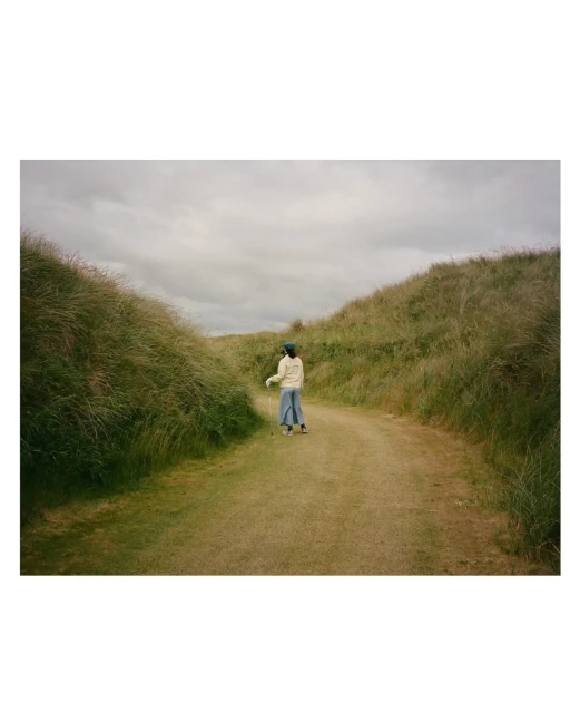 A person walks down a grassy path between tall, grassy dunes under a cloudy sky. The person is wearing light-colored clothing.