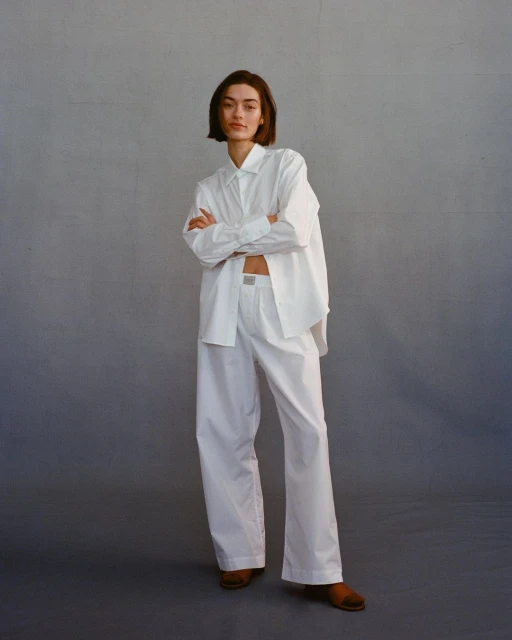 A fair-skinned woman with short brunette hair poses in a white button-down shirt, white pants, and brown shoes in a studio setting.