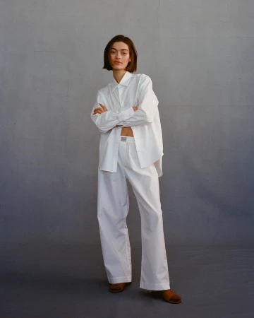 Woman with short dark hair standing with arms crossed wearing a white shirt and white trousers against a grey backdrop