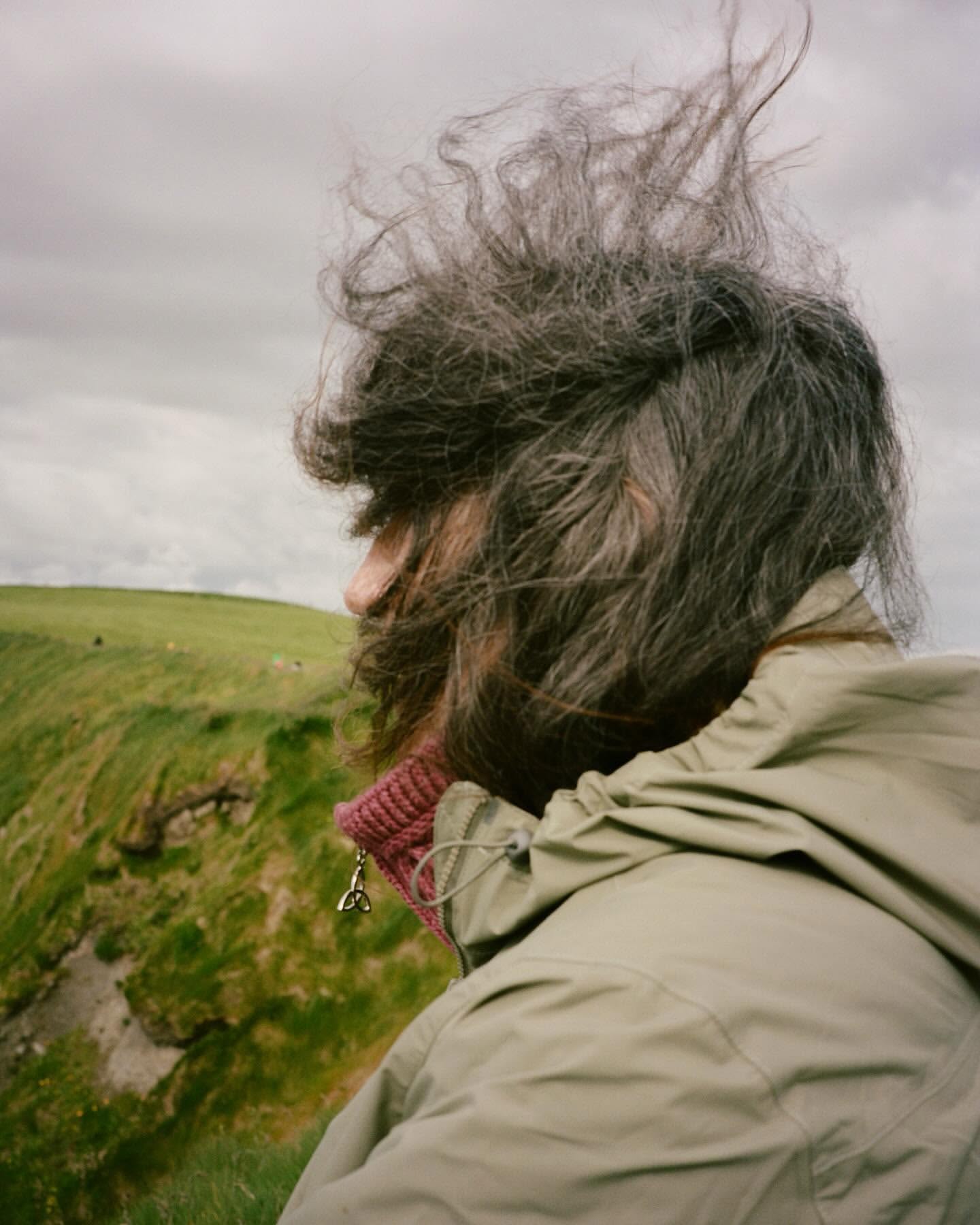 Adult with tousled hair faces left in an outdoor shot. She wears a tan jacket against a green hillside under a cloudy sky.