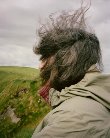 Adult with tousled hair faces left in an outdoor shot. She wears a tan jacket against a green hillside under a cloudy sky.