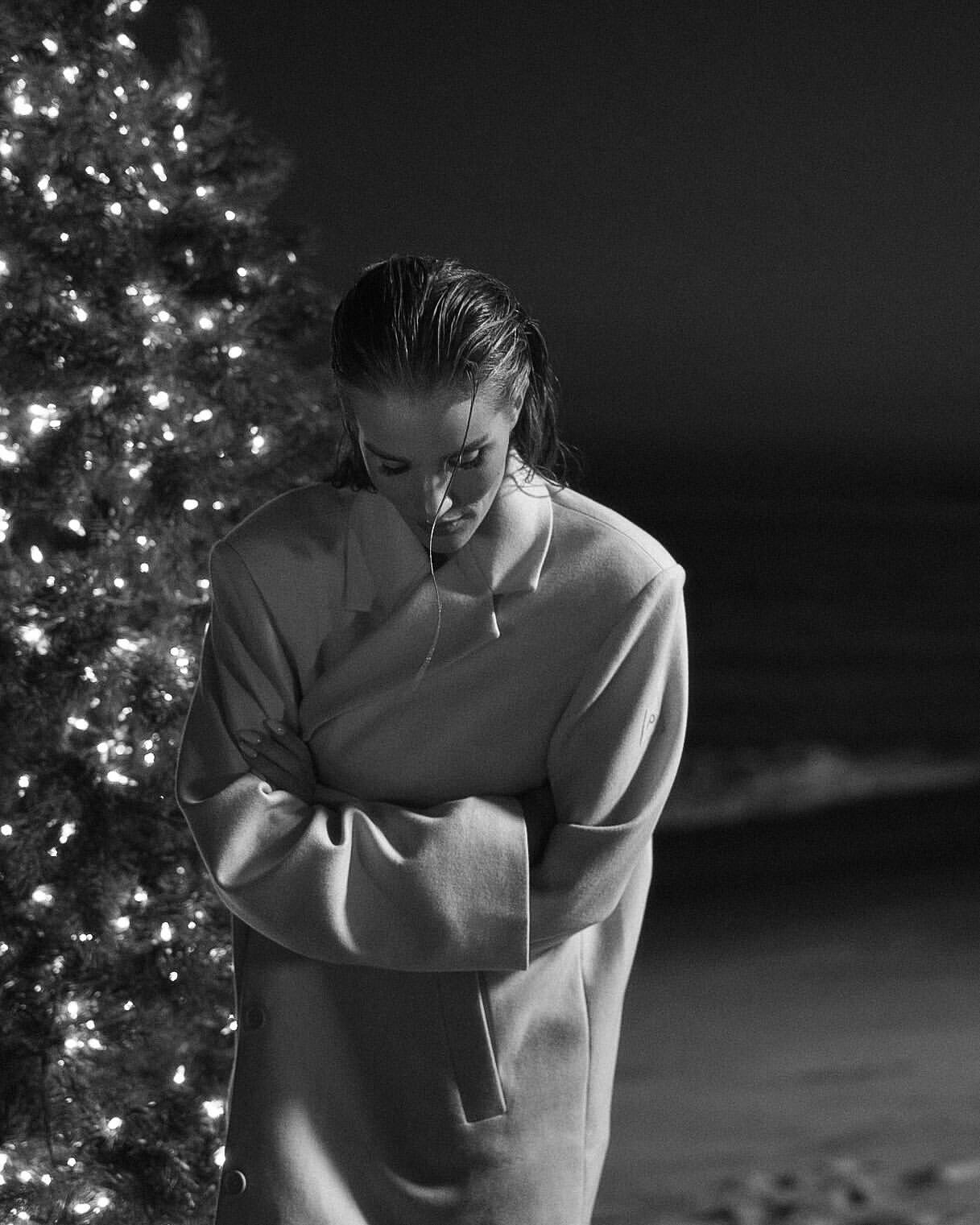 Monochrome portrait of a woman in a coat standing next to a lit Christmas tree on a beach, looking down, with crossed arms.