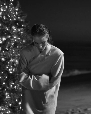 Monochrome portrait of a woman in a coat standing next to a lit Christmas tree on a beach, looking down, with crossed arms.