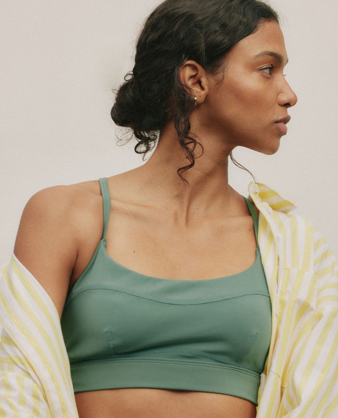 Portrait of a woman in a green sports bra and yellow striped shirt, looking off to the side, with a neutral studio background.