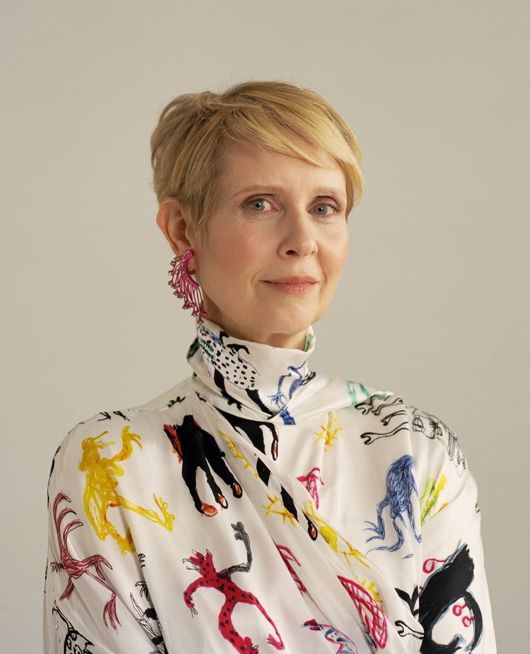 Cynthia Nixon portrait. Short blonde hair, wearing a white patterned high-necked top and pink earrings, against a plain background.