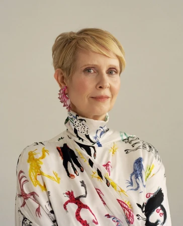 Cynthia Nixon portrait. Short blonde hair, wearing a white patterned high-necked top and pink earrings, against a plain background.