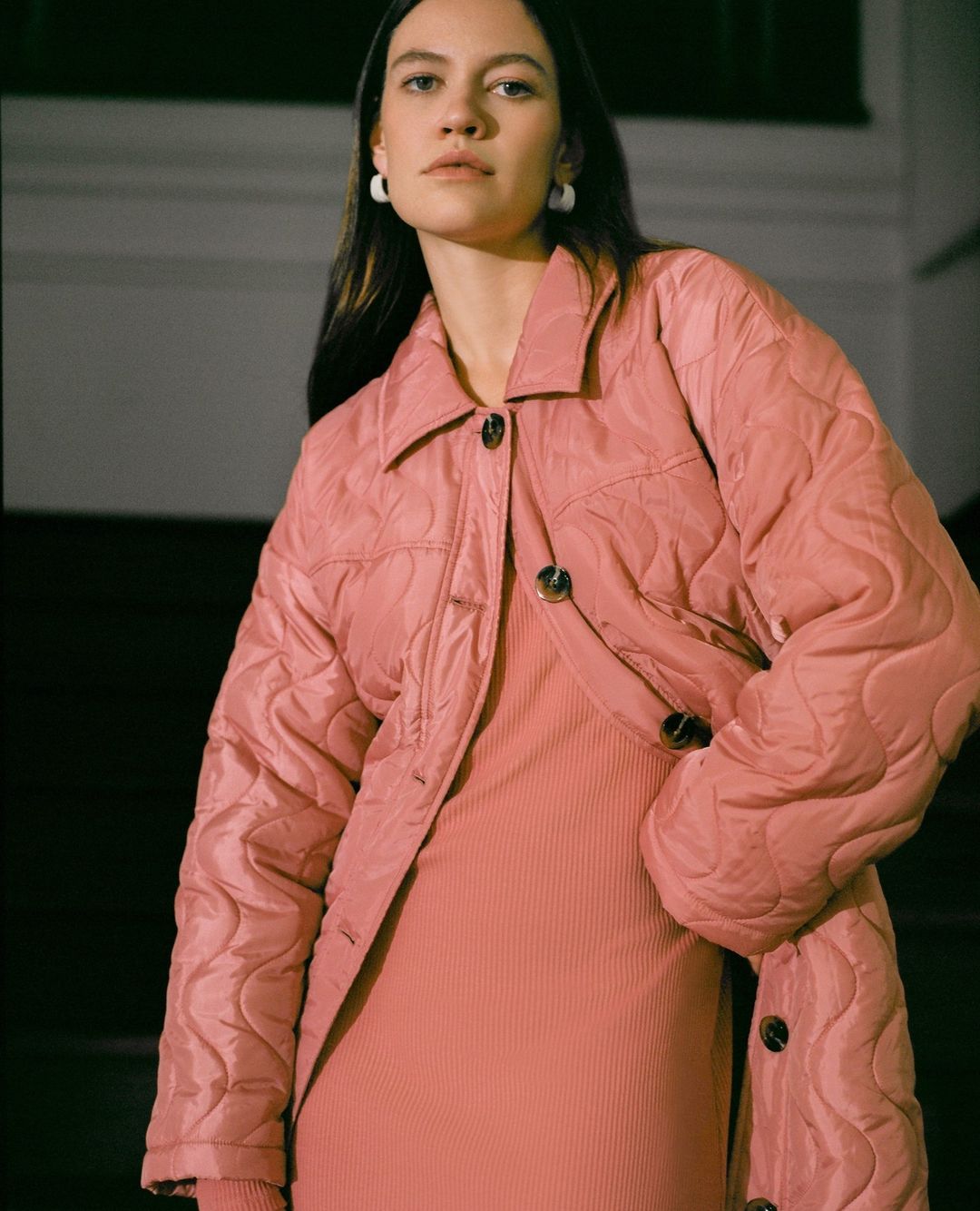 A brunette woman in a pink quilted jacket and dress stands against a dark staircase in a medium shot, showcasing a casual style.