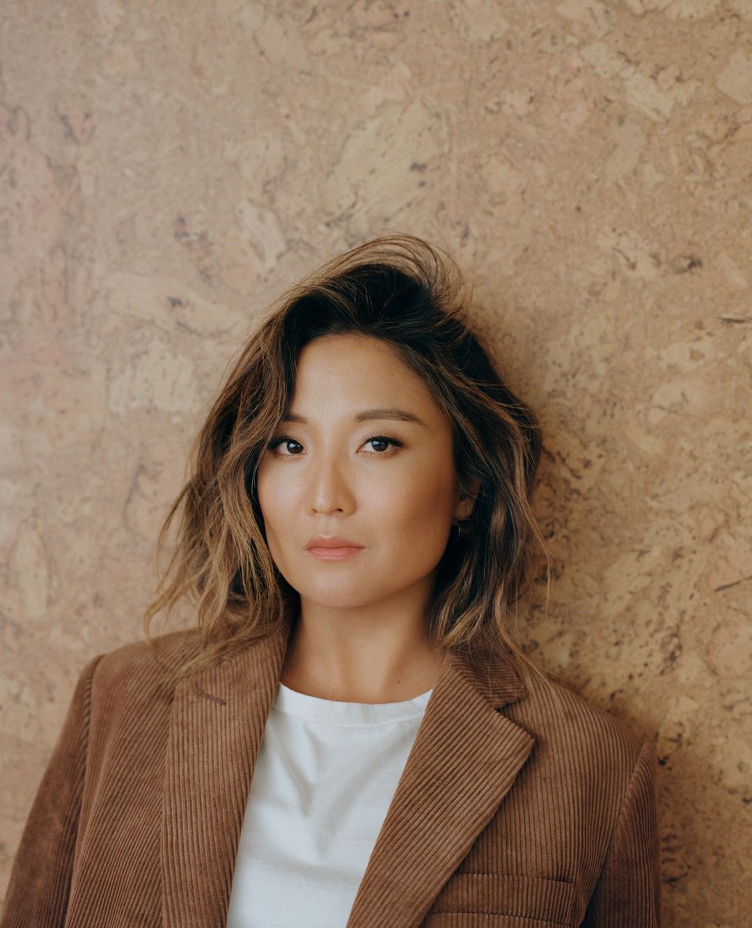 Portrait of an adult Asian woman wearing a corduroy blazer and white shirt against a textured beige wall, posing indoors.