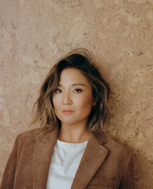 Portrait of an adult Asian woman wearing a corduroy blazer and white shirt against a textured beige wall, posing indoors.