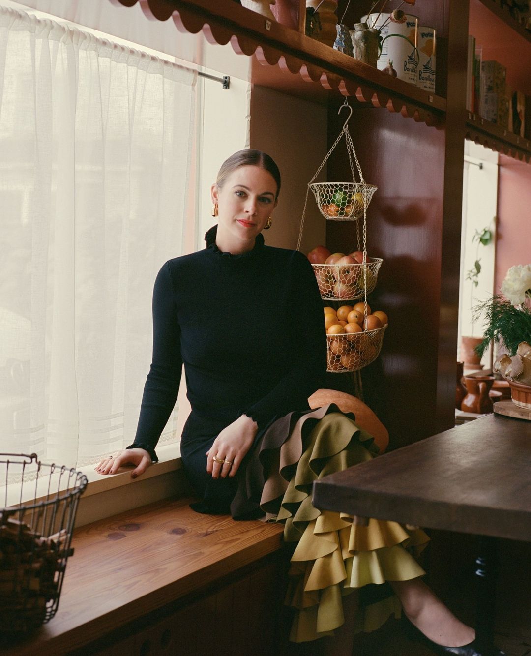 Woman in black turtleneck and tiered skirt sits on a window seat with decorative shelves, hanging fruit basket, and flowers nearby.
