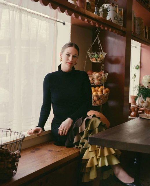 Woman in black turtleneck and tiered skirt sits on a window seat with decorative shelves, hanging fruit basket, and flowers nearby.