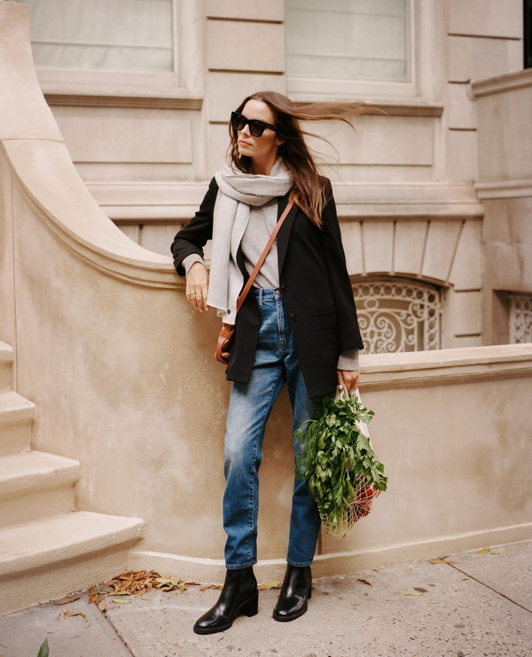 Stylish woman in black blazer, jeans, and boots stands on city street with a bag of fresh greens and brown crossbody bag.