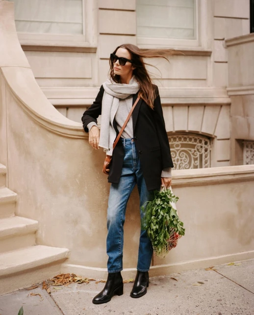 Stylish woman in black blazer, jeans, and boots stands on city street with a bag of fresh greens and brown crossbody bag.
