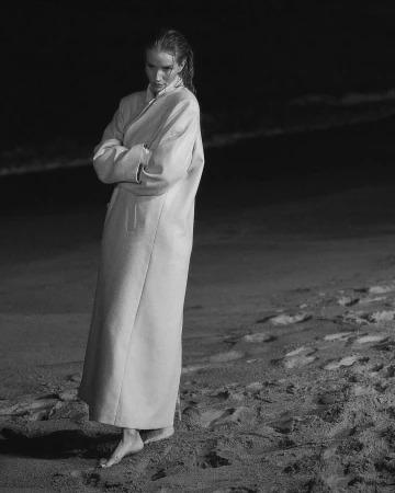 Woman in a long light-colored coat standing barefoot on a sandy beach at night.