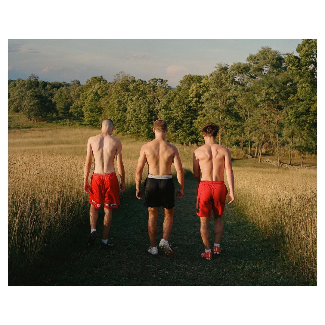 Three shirtless adult males are seen from the back, walking in a field of wheat towards a treeline under a bright, clear sky.
