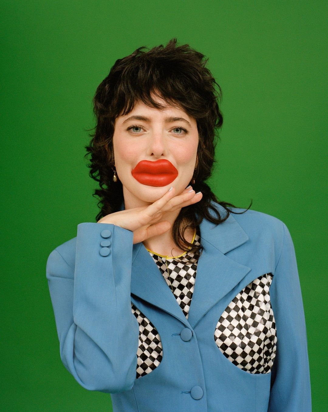 Woman with large lipstick mark on her face, wearing a blue jacket against a green background in a surreal and playful composition.