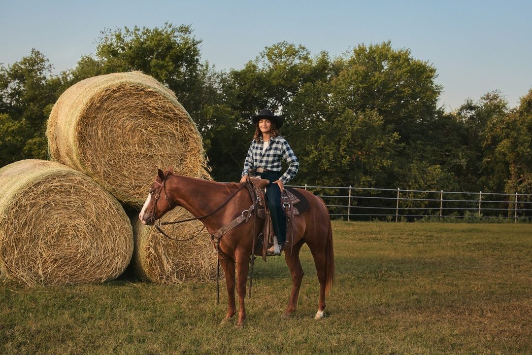 Woman in a black hat rides a chestnut horse in a grassy field next to hay bales, backed by trees under a clear sky.