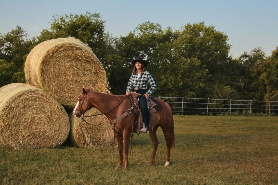 Woman in a black hat rides a chestnut horse in a grassy field next to hay bales, backed by trees under a clear sky.
