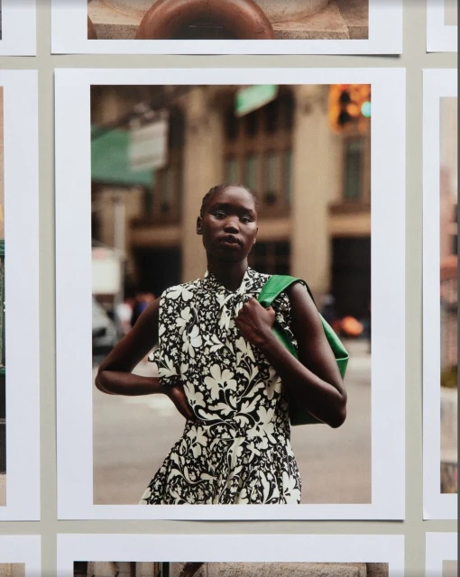 Fashion editorial shot of a Black woman in an elegant patterned dress with a green bag, standing in an urban street with buildings and traffic.