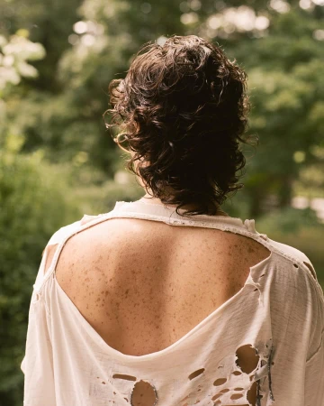 Portrait from behind of a person with curly dark hair wearing a distressed white t-shirt showing freckled back